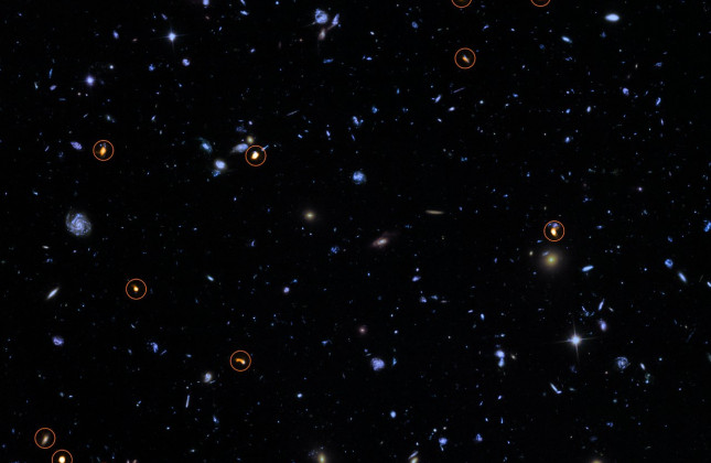 Deze foto is een combinatie van een achtergrondfoto, gemaakt door Hubble van NASA en ESA (blauw/groen) en een nieuwe, zeer diepe ALMA-opname van hetzelfde gebied (oranje en omcirkeld). Alle objecten die ALMA ziet blijken zware, sterren-vormende sterrenste