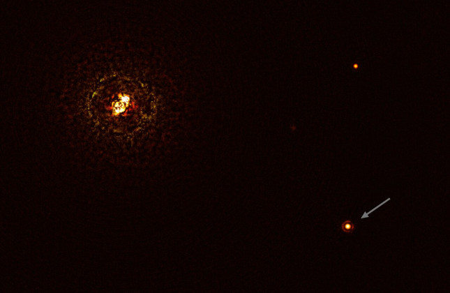 Foto van het zwaarste sterrenpaar (linksboven) met planeet (rechtsonder bij de pijl) dat tot nu toe is ontdekt. (c) ESO/Janson et al