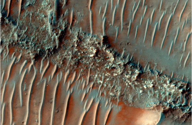 Kleurenfoto van de sterk geërodeerde stroomruggen in Noachis Terra op Mars. Her en der worden de ruggen doorkruist door zandduinen. © HiRISE Image: ESP_085386_1505 NASA/JPL/University of Arizona