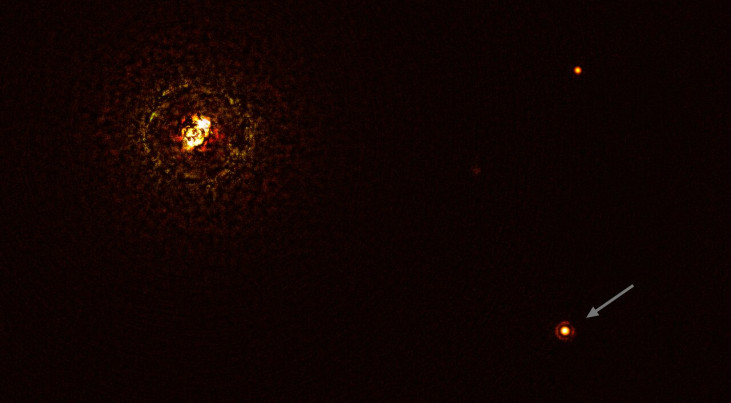 Foto van het zwaarste sterrenpaar (linksboven) met planeet (rechtsonder bij de pijl) dat tot nu toe is ontdekt. (c) ESO/Janson et al